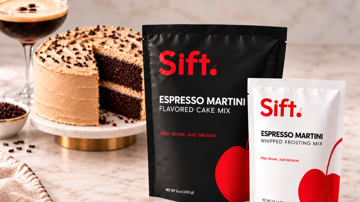  Sift Espresso Martini flavored cake mix packages, a black bag of espresso martini flavored cake mix and a white bag of espresso martini whipped frosting mix, displayed on a marble countertop next to a slice of chocolate cake and a cocktail glass.